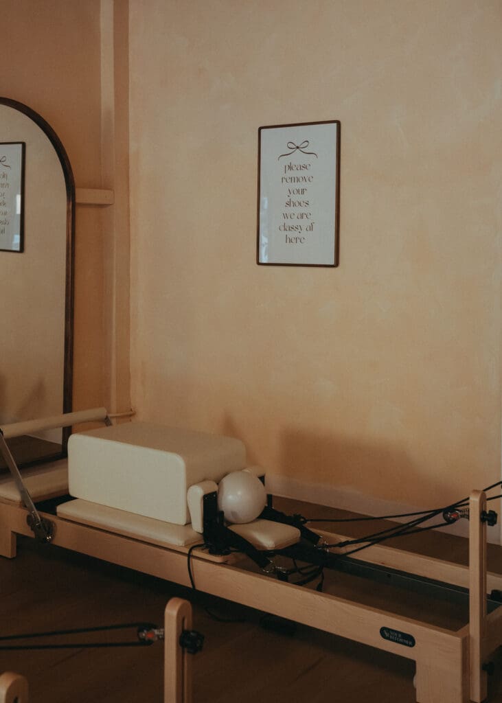 Close-up of reformer machine and studio sign reading “please remove your shoes” in wellness studio.