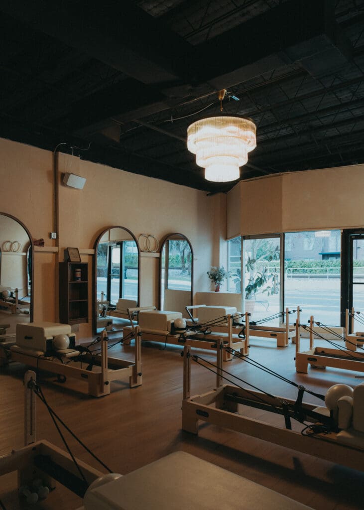 Wide interior photo of modern Pilates studio with reformers, neutral tones, and wood panel walls.