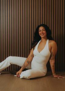 Portrait of Pilates instructor in a white set sitting on the studio floor against a wood slat wall, photographed for personal branding and fitness photography.
