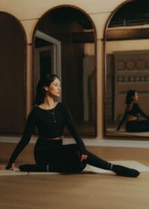 Pilates instructor seated on mat in warm, moody wellness studio.
