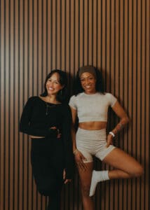 Two Pilates instructors standing against wood panel wall laughing during branding session.