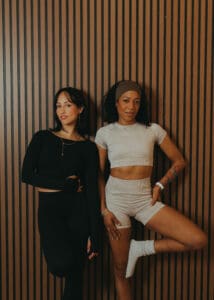 Two Pilates instructors standing against wood panel wall laughing during branding session.