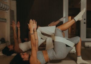 Group of women performing reformer Pilates single leg glute bridge exercise in wellness studio.