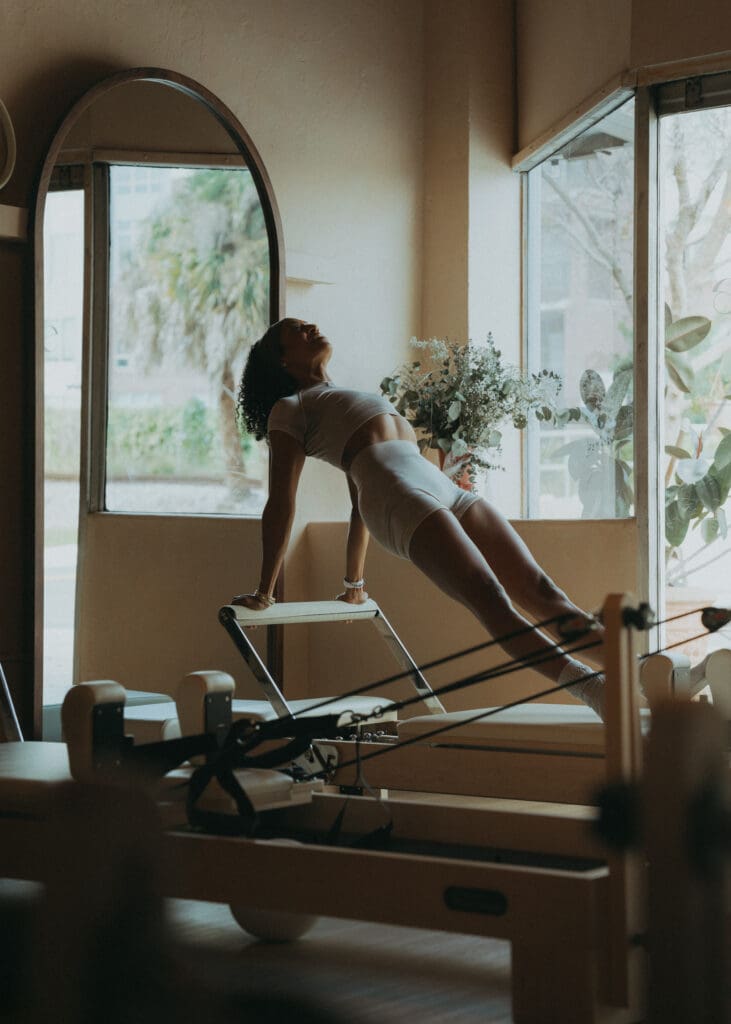 Woman performing reformer Pilates bridge exercise near large window with soft natural light.
